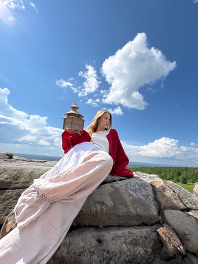 A young woman with long blonde hair sits on large rocks on a sunny day. She is wearing a white top, a red jacket, and a long, flowing pink skirt. In her hands, she holds a small wooden model of a building. The background shows a bright blue sky with fluffy white clouds and a distant green forest. The location is the Kuberla rock formation, also known as the "Devil's Finger," in Gubakha, Russia, a popular tourist attraction and natural landmark.