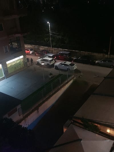 This nighttime image captures a quiet urban street scene in Tiranë, Albania, viewed from an elevated perspective, likely from a window or balcony overlooking the road. The sky is dark, indicating the late evening or night.
A row of cars is parked along one side of the street, extending into the distance. Prominently featured are a white sedan in the foreground and a red car behind it, along with several other darker vehicles further down the street. The scene is illuminated by artificial light sources, including a bright, modern streetlight on the left side of the frame, which casts light on the parked cars and the road surface. Some car headlights are also visible.
On the right side of the street, a multi-story building with distinct architectural features, possibly balconies or window recesses, is partially visible. Another building is vaguely discernable on the far left. The road itself appears paved, with a curb separating it from the parked vehicles. Patches of foliage or trees are visible in the lower right corner and in the background, though details are obscured by darkness.
No specific human activity or event is taking place; the street appears calm and static. The weather seems clear, with no rain or other atmospheric conditions evident. There is no visible text in the image.