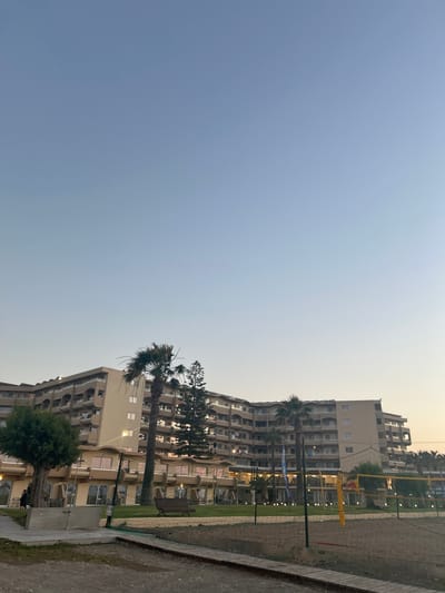 This is a vertically oriented, outdoor, eye-level photograph of a resort complex, likely in Rhodes, Greece. The sky takes up the majority of the left side of the frame and is a pale blue, transitioning to a light yellow near the horizon on the right, indicating it is either dawn or dusk.
The primary subjects are several multi-story resort buildings with light-colored facades. These buildings appear to be modern hotels or apartment complexes, with numerous balconies and windows visible. A few palm trees and other lush greenery are interspersed around the buildings and in manicured lawns.
In the foreground on the right side of the image, a sandy area is visible, suggesting a sports court, possibly for beach volleyball, as indicated by a net and yellow poles. A paved walkway runs alongside this area.
There are no clearly discernible people engaged in any specific activity, though a few figures might be present in the lower parts of the buildings, too indistinct to be sure. The overall mood is peaceful and serene, enhanced by the soft, natural light of the transitional time of day.
No visible text is present in the image. The scene depicts a tranquil resort setting in a coastal or near-coastal location, likely a tourist destination given the architecture and amenities.