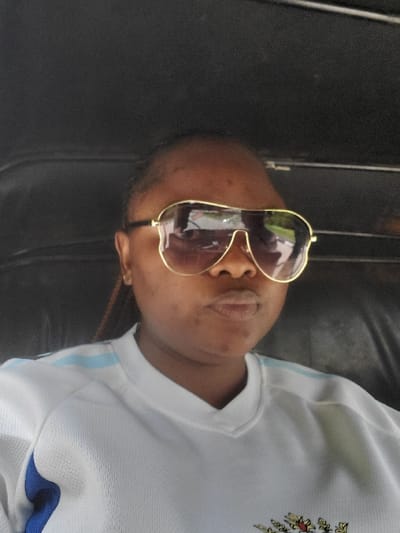 An adult female is depicted in a close-up, head-and-shoulders shot. Her complexion is dark, and her hair appears to be styled in braids. She wears large, aviator-style sunglasses with gold-colored frames, reflecting ambient outdoor light and partial views of what appears to be foliage or sky. Her mouth is slightly pursed, and her gaze is directed forward, likely at the camera. She is wearing a white short-sleeved shirt with light blue stripes visible on the shoulders and a blue panel under the arm. A crest or logo, possibly embroidered, is partially visible on the lower front of the shirt. The background is dark and indistinct, suggesting an enclosed interior space, possibly a vehicle, with dark textured upholstery visible. There is no visual information within the image to confirm the location as Isunba, Nigeria.