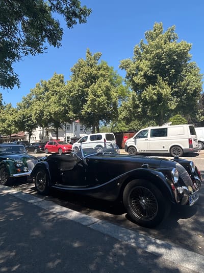 This is a medium shot, eye-level, outdoor photograph taken on a sunny day. The scene depicts a street lined with parked cars and trees. In the foreground, two classic cars are prominently featured. The car closest to the viewer is black with a visible wire-spoke wheel and a license plate that appears to read "HN MO 102". To its left is a dark green classic car with a shiny grille. Further down the street, other vehicles are visible, including a red hatchback, a white van, and another white van. Buildings, likely residential or commercial, are present in the background, partially obscured by lush green trees. The sky is a clear, vibrant blue. The location is identified as Heilbronn, Germany, specifically on Wilhelm-Leuschner-Straße, near the intersection with Allee. The street is paved, and a curb is visible. Shadows from the trees are cast across the pavement. There are no people visible in the image.