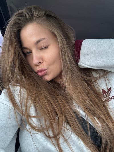 A young woman with long, brown hair is captured in a selfie, winking and puckering her lips. She is wearing a light grey hoodie with a dark red trim and an Adidas logo on the chest. Her hair is slightly tousled and falls across her face and shoulders. The background is dark and out of focus, suggesting an indoor setting, possibly a car. There is no indication of the city of Kueda, Russia, in the image.