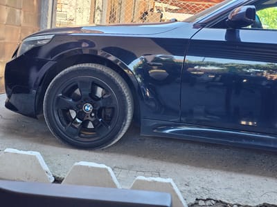 The image shows a close-up, upside-down view of the side and wheel of a dark blue BMW. The car appears to be parked outdoors on a concrete surface, with a cracked asphalt or concrete area visible in the background above the car. There are some white, angular concrete blocks or structures also in the background, casting shadows. The lighting suggests it is daytime, likely sunny, as evidenced by the reflections on the car's polished surface. The reflection shows what appears to be greenery and a structure with horizontal lines, possibly a building or fence.
The wheel is black and has the BMW logo in the center. The car's side panel, door handle, and side mirror are also visible. The setting appears to be a residential or possibly a service area, with a chain-link fence and some brickwork visible at the bottom of the frame. No people are visible in the image. The overall scene is static, with no discernible activity taking place. The image does not contain any visible text. The location context is Skutare, Bulgaria.
