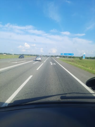 A forward-facing, first-person perspective from inside a vehicle on a highway shows a clear blue sky with scattered white clouds. The road has two lanes in the direction of travel, with white lane markings. A silver car is in the right lane, and a dark SUV is in the left lane. Ahead, a blue road sign indicates "ВАГОННИК-2 3" with arrows pointing to the right, suggesting an upcoming exit or turnoff. To the right of the road, a grassy shoulder leads to a line of trees and a blue and white sign, possibly for a gas station, with Cyrillic text and prices. The foreground shows the dashboard and windshield wipers of the vehicle. The scene appears to be on a highway near Pokrovka, Russia, based on the road sign.