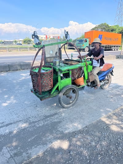 In Pamotan, Indonesia, a man wearing a straw hat and a dark t-shirt rides a three-wheeled motorcycle with a green wooden cart attached. The cart is equipped with a large bamboo basket filled with green items, a metal container, and an orange bucket. Two decorative figures, one resembling a puppet and the other a butterfly, are attached to the front of the cart. In the background, a large orange Hapag-Lloyd shipping container truck drives on a highway, with lush green trees and buildings visible on the side. The sky is clear blue with fluffy white clouds. The motorcycle and cart are on a paved road with patches of gravel and debris.