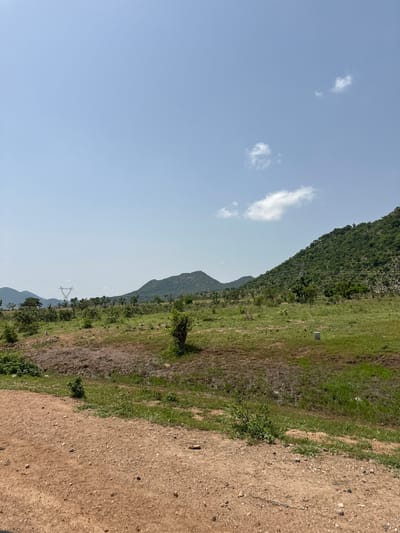 The image depicts a rural landscape under a clear blue sky with a few scattered clouds. In the foreground, a dirt road or path with light brown soil and scattered small stones leads into the scene. Beyond the road, a grassy field stretches out, interrupted by patches of dry, muddy earth. Further into the background, a line of trees and vegetation separates the field from rolling hills. Two prominent hills, covered in dense green foliage, rise in the mid-ground and background, with one hill dominating the right side of the frame and another further to the left. A large electrical pylon, a common sight in rural areas for power transmission, stands between the hills in the middle distance. The overall impression is one of a natural, undeveloped environment. There are no people, animals, or man-made structures, apart from the pylon, visible in the image. The image is a high-angle shot, looking slightly down towards the landscape.