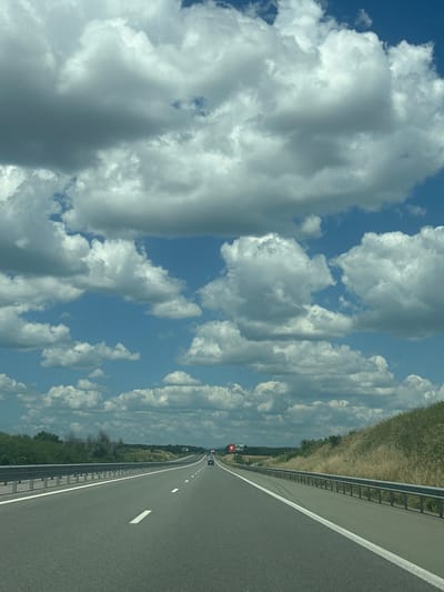 The image shows a straight, paved highway leading into the distance. The highway has two lanes in each direction, separated by a solid white line. There are white dashed lines marking the lanes within each direction of travel. On the left side of the highway, there is a metal guardrail with posts. Beyond the guardrail, there is a grassy embankment with some bushes and trees. On the right side of the highway, there is also a grassy embankment with dry grasses. The sky is blue with numerous large, white, fluffy clouds. In the distance, there are a few cars on the highway, and a billboard can be seen on the left side. The overall impression is of a typical highway scene on a bright, partly cloudy day. The location is identified as the highway near Chirpan, Bulgaria.
