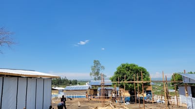 This wide outdoor shot captures a bustling construction or development site in Tchere, Democratic Republic of the Congo, under a vast, clear blue sky dotted with a few small white clouds. The bright daylight suggests midday or early afternoon.

In the foreground, the ground is raw earth, indicative of ongoing work, with wooden planks and beams scattered about as building materials. To the left, a newly constructed building with white fabric or tarp walls and a corrugated silver metal roof stands prominently. Two individuals in dark clothing are seen bending over, actively engaged in work near its base. Throughout the mid-ground, several small, single-story buildings are visible; some feature yellowish wooden walls and blue corrugated metal roofs, with what appear to be solar panels installed on some. White fencing or railings are also present around these structures. Another person in a light top is visible near one of the yellow buildings.

Wooden scaffolding and upright poles are interspersed across the site, contributing to the sense of active construction. Lush green trees punctuate the landscape, with a large, full tree on the right and a tall, slender tree in the mid-background. Bare tree branches frame the top-left corner. In the distant background, green hills and mountains stretch under the horizon, suggesting a rural or semi-rural setting.

The image portrays a scene of diligent work and community progress. A watermark "Joseph Kisuki" is visible in the lower right corner.