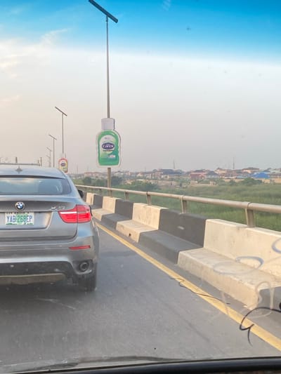 On a bridge with alternating black and white barriers, a grey BMW X5 travels on the road. A yellow line separates the traffic lanes. Advertisements for Carex hand sanitizer are positioned on poles along the roadside. The license plate reads “YAB-268EP”, and identifies the car as registered in Abuja. A faint cityscape stretches along the horizon under a hazy sky. Part of the window’s frame and some reflections are visible in the foreground.