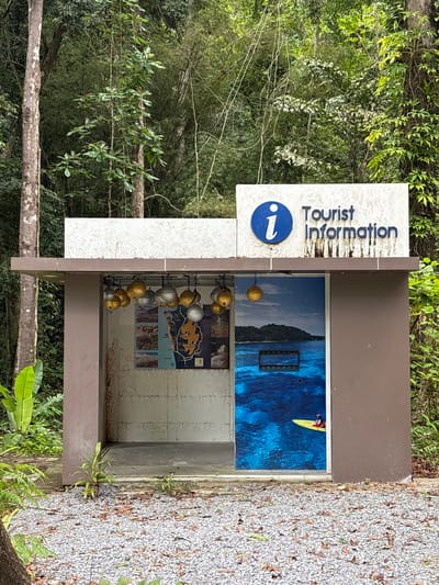 A tourist information kiosk stands in Tambon Thai Mueang, Thailand, surrounded by lush foliage. The kiosk is constructed of brown and white materials with a sign on top that reads "Tourist Information" next to an "i" symbol. Inside the kiosk are various informational displays, including images of a beach scene with a person on a yellow kayak, a map of the area, and other related pictures. A string of gold and silver spheres hangs within the kiosk. The ground around the kiosk is covered in small gravel and foliage. Trees and dense greenery fill the background.