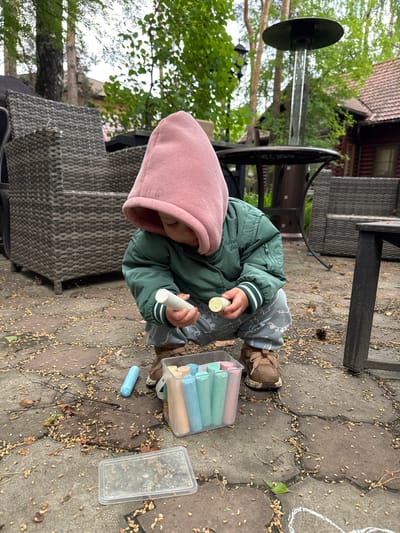 A young child wearing a pink hooded jacket and a green jacket sits on a paved surface surrounded by fallen leaves and debris. The child holds chalk pieces in both hands. A plastic container next to the child holds more chalk pieces. To the left, there is a patio table and wicker chairs. The setting appears to be outdoors, possibly in Tomilino, Russia. The child is engaged in playing with the chalk, likely drawing on the pavement.