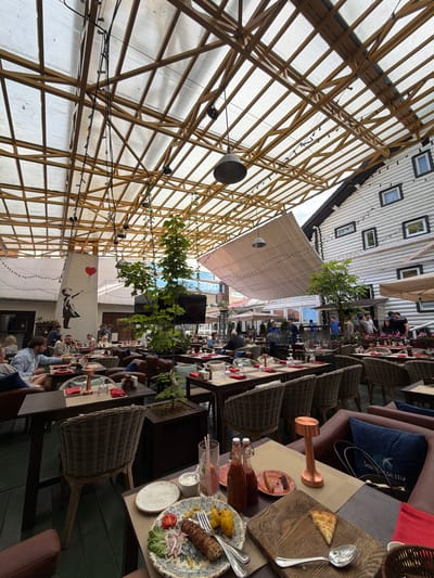 The image depicts a spacious outdoor seating area of a restaurant, likely located in Zhukovka, Russia, characterized by a wooden-framed, transparent roof structure and a rustic, log-cabin-style building facade. The area is filled with tables and chairs, some occupied by patrons.

In the foreground, a table is set with a meal, including a kebab, fries, and a slice of pizza on a wooden board. A spoon and fork are also visible on the table, along with two bottles of sauce and a small plate of olives. A glass with a drink and ice is present, as is a small bowl of dipping sauce. A copper-colored table lamp stands nearby.

The background shows a lively scene with various groups of people dining and socializing. The restaurant's interior, visible through windows and open doorways, appears to be furnished in a similar rustic style. The overall atmosphere suggests a well-established restaurant catering to a relaxed, upscale clientele, with a focus on outdoor dining during daylight hours. The architectural style, particularly the log building and the expansive wooden roof structure, strongly suggests the location is within a dacha or countryside setting, consistent with Zhukovka's reputation as an affluent rural district outside Moscow.
