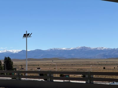 The image captures an expansive outdoor landscape under a clear, bright blue sky, likely taken from an elevated vantage point such as a bridge or overpass.

In the immediate foreground, a dark gray concrete structure spans the bottom of the frame, topped by a lighter-colored, multi-barred wooden or composite fence that stretches horizontally across the lower third of the image. The fence has several vertical posts. To the right, below the fence line, a white car, possibly a sedan or small SUV, is partially visible, driving on a road from right to left. A small green sign with white text, reading "Salida" (Spanish for "Exit"), is partially visible to the left of the car, just below the fence.

Dominating the left side of the foreground and extending into the midground is a tall, slender white pole. Mounted near the top of this pole are several dark, rectangular fixtures, which appear to be a combination of solar panels and/or light fixtures, oriented at various angles. A dark green conifer tree, possibly a pine, is visible behind the fence and to the left of this prominent pole. Further into the midground, behind the fence, another, smaller light-colored pole with what appears to be a spherical light fixture is visible on the right.

The midground unfolds into a vast, open, and relatively flat landscape characterized by dry, light brown and tan earth. This terrain suggests a steppe or high-desert environment, with sparse, low-lying vegetation scattered across the ground. The land gently rolls into low hills in the distance.

In the background, a long and impressive range of mountains stretches across the entire horizon. The peaks and upper slopes of these mountains are covered in significant patches of white snow, indicating a high elevation or cold climate. The lower slopes of the mountains appear darker, possibly forested or rocky. The sky above is a pristine, cloudless blue, suggesting a sunny day with excellent visibility.

The overall impression is one of wide-open spaces and natural beauty, juxtaposed with elements of modern infrastructure. The time of day appears to be midday or early afternoon, given the bright, overhead lighting and absence of long shadows.