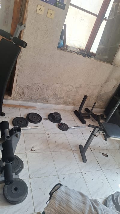 The image depicts an interior scene, likely a room with a dirt floor covered in white tiles. The room appears to be a storage or workshop area. Visible objects include a weight bench with black padding, several weight plates of various sizes scattered on the floor, and barbell equipment. In the background, there is a rough, unplastered wall with two light switches. A window with a wooden frame and translucent covering is also present. The overall impression is one of disarray and neglect, suggesting the room is not actively maintained or used for its intended purpose. The location of Lom, Bulgaria, is not discernible from the image itself.