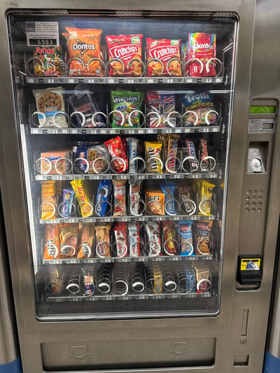 The image shows a vending machine filled with various snacks. The vending machine is located in Duisburg, Germany, as indicated by the text on a label at the top left which reads "Im Gartenkamp 110-112, 49492 Westerkappeln" and a phone number that begins with "05404". The machine has multiple rows of snacks, including chips, cookies, candy bars, and gummies. Each item is priced in Euros and has a corresponding number for selection. The machine's front is made of glass, allowing the contents to be clearly visible. A coin slot and a bill acceptor are visible on the right side of the machine, along with a keypad for product selection. The environment appears to be indoors, possibly a public space given the nature of a vending machine. There are no people visible in the image.