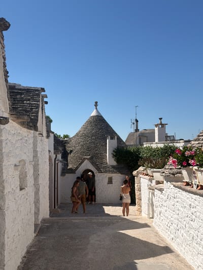 In Alberobello, Italy, a narrow, paved alleyway winds between traditional white trulli houses with conical stone roofs. The sun casts shadows on the ground. Several people are visible. On the left, two women are walking away from the camera. In the center, a group of three people, two men and a woman, are gathered near an entrance to a trullo. One of the men appears to be wearing a white cap. Further into the background, another trullo with a distinctive finial on its roof is prominent. On the right side of the alley, a white wall adorned with potted plants, including pink flowers and succulents, lines the pathway. The sky is a clear, bright blue.