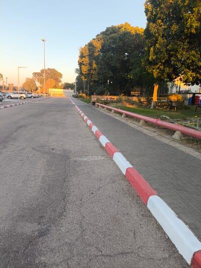 The image shows an outdoor scene on a paved road in Dimona, Israel. The road is lined with a sidewalk constructed of interlocking bricks. A red and white striped curb separates the road from the sidewalk. On the right side of the sidewalk, a red pipe is elevated on concrete supports. Beyond the sidewalk, there are trees, some with illuminated leaves due to the setting sun. In the background, a building with a yellow awning and a parking lot with several cars can be seen. The sky is clear and blue, indicating it is daytime, likely late afternoon or early evening given the lighting. There are no people visible in the image.