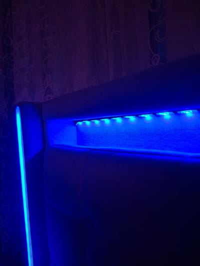 The image shows a close-up view of a headboard with integrated blue LED lighting. The headboard appears to be upholstered in a light-colored fabric. A vertical strip of bright blue light is visible on the left side of the headboard, and a horizontal strip of blue LEDs illuminates a recessed area within the headboard. The background consists of dark, possibly purple-tinted curtains with a subtle patterned texture. The lighting creates a moody and atmospheric effect. The image is taken indoors, likely in a bedroom. There is no indication of a specific location within the city of Evere, Belgium.
