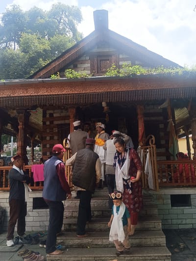 The image shows a group of people gathered outside a wooden structure, possibly a temple, with intricate carvings. Several individuals are wearing traditional attire, including caps and vests. Some are ascending a set of stone stairs leading into the building. A young child in traditional clothing is also present on the stairs. The background features lush green trees and a cloudy sky. The overall atmosphere appears to be that of a cultural or religious gathering. The location is not identifiable as Gojra, India.