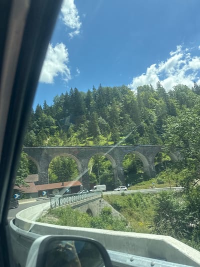 A wide, sunlit outdoor shot captures a scenic mountain landscape in Hauts de Bienne, France. A large stone viaduct with multiple arches dominates the mid-ground, spanning across a valley. A paved road curves in the foreground, bordered by a concrete barrier and a guardrail, with a small bridge extending from it. Cars are visible on the road, including a white SUV and a van. Lush green trees and vegetation cover the steep slopes of the mountain, reaching towards a clear blue sky dotted with white clouds. A building with a reddish-brown roof is partially visible behind the viaduct on the left. The view appears to be from inside a moving vehicle, as evidenced by the reflection in the side mirror and the dark frame of the vehicle's interior on the left side of the image.