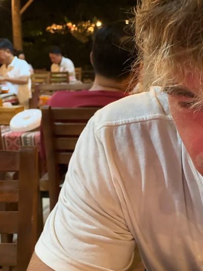 This image depicts a scene at an outdoor restaurant in Tu Le, Vietnam, likely during the evening. The focus is on the right shoulder and upper arm of a person wearing a white t-shirt. Their blonde hair is visible in the foreground, obscuring part of their face. In the background, blurred figures are seated at tables. To the left, a person in a white polo shirt is visible. Another person in a red shirt is also in the background. Wooden chairs and tables are present, with some patterned fabric on a table. The lighting suggests it is nighttime, with some illuminated string lights in the distance.