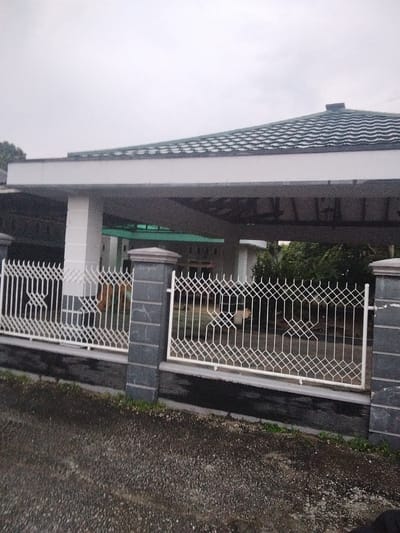 The image shows a white metal fence with a geometric pattern in front of a building. The building has a white facade and a green tiled roof. The sky is overcast. The ground in front of the fence is paved with gravel. The scene appears to be located in a residential area.