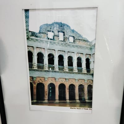 This media file is a photograph, likely framed, depicting a historic building. The main subject is an ornate, multi-storied structure, possibly a house or palace, exhibiting classical architectural influences with elements of decay.
The building features a series of arched openings on both visible floors. The lower floor presents several large, dark arched doorways or windows, set into walls of a light reddish-brown or peach hue, adorned with intricate white or light-colored decorative carvings above each arch. This floor is reflected in a calm body of water, possibly a pond or canal, in the immediate foreground.
The upper floor mirrors the lower's arched design, featuring balconies with dark railings, supported by numerous white fluted pillars between the arches. The decorative elements, including floral and scrollwork patterns, are consistently seen on both levels. The uppermost part of the building appears to be in a state of ruin or disrepair, with exposed brickwork and crumbling sections, while still retaining some of its original intricate ornamentation. The overall color palette is soft, with light tones dominating the sky and the building's decorative accents, suggesting a bright or overcast day.
No people or specific activities are visible in the image. The photo evokes a sense of historical grandeur and the passage of time.
Visible text at the bottom right corner of the photograph clearly states: "Niharika House in Panam City".