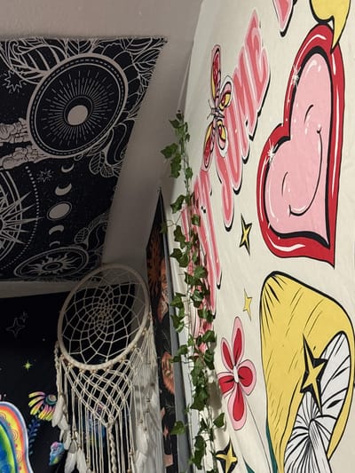 A wall tapestry depicting various colorful, cartoon-like images is prominently displayed. The tapestry features a large pink heart with a smiling face, a yellow mushroom with a star inside, and pink flowers. A string of artificial ivy is draped along the tapestry. To the left, another tapestry with a celestial, black and white sun and moon design is visible. A white dreamcatcher with feathers hangs from the top of the frame, partially obscuring a black tapestry with colorful celestial designs. The overall scene appears to be indoors, likely a bedroom, with a focus on decorative wall hangings. The location of this scene within Gaylord, United States of America, cannot be determined from the provided image.