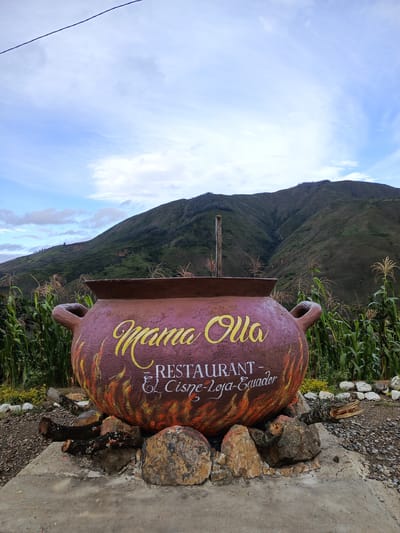 A photograph captures a large, pot-shaped sign in the foreground with the words "Mama Olla" in yellow script, and "Restaurant - El Cisne-Loja-Ecuador" in white. The pot is brown with painted flames at the bottom. It rests on rocks and logs on a concrete slab. Behind the sign, green corn stalks grow on either side. In the background, a lush green mountain slopes upward under a sky with scattered clouds. The scene is set in El Cisne, Loja, Ecuador, as indicated on the sign.