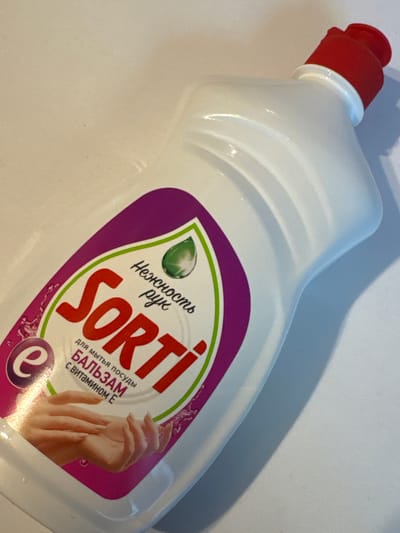 A white plastic bottle with a red cap is shown at an angle against a white background. The bottle has a purple and green label with text and an illustration of hands. The text on the label includes "Sorti", "Нежность рук" (Tenderness of hands), "для мытья посуды" (for washing dishes), and "БАЛЬЗАМ С ВИТАМИНОМ Е" (Balm with vitamin E). The label also features a green droplet graphic and a graphic of two hands. The bottle is illuminated from the side, casting a shadow to the right. There are no people visible in the image. The environment is a plain white surface. No specific location in Яромінскі сельскі Савет, Belarus can be identified from this image.