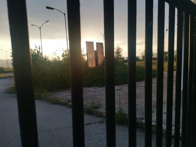 The image captures an outdoor scene in the city of Zhivkovo, Bulgaria, viewed through the dark, vertical bars of a fence. Approximately ten to twelve dark, evenly spaced vertical bars are prominent in the immediate foreground, partially obstructing the view. The scene is illuminated by the warm, low light of a sunrise or sunset, emanating from the horizon.
In the midground, an asphalt road, visibly cracked and overgrown with weeds, transitions to a paved area composed of square or rectangular tiles, which also exhibit significant weed growth. Beyond this, a dense cluster of low-lying green bushes extends across the center of the frame. Two prominent, tall, rectangular concrete slabs or monuments stand centrally, directly back-lit by the sun, creating strong silhouettes. The left slab features visible markings or discoloration on its surface.
Numerous streetlights with single or double lamps are scattered throughout the background, their dark forms silhouetted against the bright, hazy sky which progresses from a warm orange/yellow glow near the horizon to a lighter blue/grey higher up. Further in the background, sparse trees and vegetation are visible, with distant hills or mountains forming the horizon on the left side of the frame, where power lines or towers are also faintly discernible. No individuals are present in the scene. The environment suggests a neglected or semi-abandoned area.