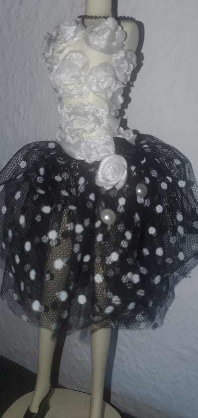This is a close-up, low-angle photograph of a mannequin wearing a doll-sized dress. The dress has a white bodice decorated with fabric roses and lace trim, and a black, polka-dotted, sheer tulle skirt. Two small white pearls are attached to the skirt. The mannequin's legs and feet are visible, with one foot wearing a dark brown shoe. The background is a textured white wall. The image does not contain information about the city of Sasbach, Germany.