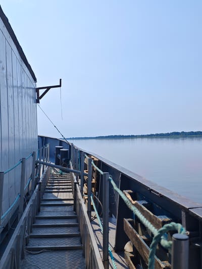 This is a medium-angle, eye-level shot taken from the deck of a vessel on a body of water. The perspective is looking down a set of metal stairs on the port side of the vessel. The stairs are flanked by metal railings with blue ropes. The side of the vessel, a large, gray, corrugated metal structure, occupies the left side of the frame. To the right of the stairs, the dark metal edge of the vessel's deck is visible, with wooden supports and a blue rope. Beyond the deck, a calm, wide body of water stretches to the horizon. On the horizon, a thin strip of lush green trees marks the distant shoreline under a clear, pale blue sky. There are no people or animals visible in the image. The scene suggests a quiet, possibly industrial or transport-related setting on a clear day. The location is identified as Novonazimovo, Russia.
