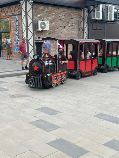 This media file features a miniature train with several carriages, predominantly red and green, with one carriage adorned with a Russian flag. Several people, including children and adults, are visible within the train's compartments. The train is positioned on a paved walkway, with a brick wall and an air conditioning unit in the background. The scene appears to be outdoors during daylight, with the weather seeming pleasant. The overall atmosphere suggests a leisurely outing or a tourist attraction.
Visible text on a building in the background reads "АПТЕКА" which translates to "APTEKA" or "PHARMACY" in Russian and Ukrainian. This suggests the location might be in an area where these languages are spoken, which aligns with the provided location context of Мысовское сельское поселение, Ukraine. The paving stones are arranged in a diamond pattern, with alternating light and dark grey colors. The train itself is detailed with a prominent red star on the front. One of the people in the foreground is wearing a maroon t-shirt and grey shorts, while another is carrying a child. The people in the train appear relaxed and engaged in the activity.