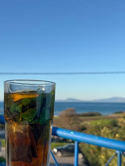 A glass of herbal tea, possibly mint, rests on a surface, juxtaposed against a bright blue sky and distant ocean. A tea bag steeps inside, alongside visible mint leaves. A blue metal railing is partially visible near the glass, hinting at a balcony or outdoor setting. Verdant foliage blurs in the background, suggesting a coastal landscape or garden. The overall scene evokes a sense of tranquility and relaxation.