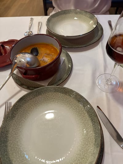 The image depicts a table setting at a restaurant. A white tablecloth covers the table, and various tableware items are arranged. In the foreground, a large white plate with a speckled grey rim is positioned, with a silver knife to its right. To the left of this plate, a fork is visible. Behind these, a small red pot filled with orange soup and a black olive sits on a grey speckled saucer. A silver ladle rests inside the pot. Further back, another grey speckled bowl with a white interior is placed on a saucer. To the right, a glass of dark red liquid, likely wine, is situated. The background is blurred, but hints of wooden furniture and a person wearing a white garment suggest an indoor dining environment. The overall scene conveys a sense of a meal being served or about to be consumed. The specific location of Gijón, Spain, is not discernible from the visual elements in the image.