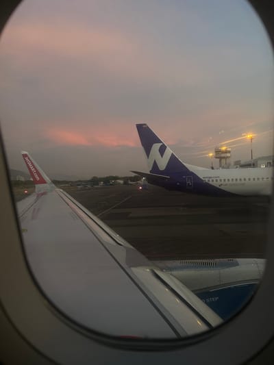 The photo was taken from the window of an airplane on the tarmac at Simón Bolívar International Airport (IATA: SMR) in Santa Marta, Colombia (latitude: 11.118, longitude: -74.232). The photo shows the wing of the plane, with the tail of another plane, a Wingo Airbus A320, visible on the right. The plane is parked on the tarmac with its tail facing the camera. The Wingo plane’s registration number, HP-1335 CME, is visible. The sky is a soft pink and grey, with the sun setting in the distance. There are some lights visible in the distance, and a control tower can be seen in the distance. The air is clear and the ground is paved.