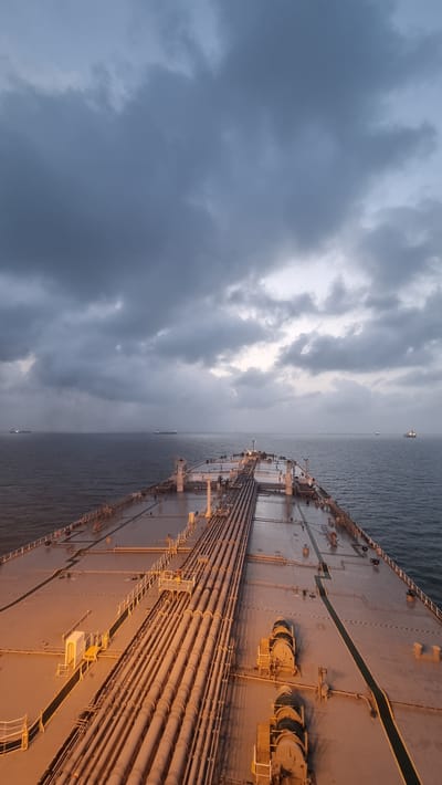 The image captures a view from the deck of a large vessel, likely a tanker, at sea. The sky is overcast with heavy, dark clouds dominating the upper portion of the frame, suggesting an approaching storm or twilight conditions. The sea is a deep, dark blue, with calm water indicating a lack of strong waves. Several other ships are visible as distant silhouettes on the horizon, scattered across the vast expanse of the ocean.

The foreground of the image shows the deck of the vessel, which is a light gray color. A prominent network of large, parallel pipes runs down the center of the deck, illuminated by a warm, golden light, possibly from the vessel's own lighting system. There are also various pieces of industrial equipment, such as winches and what appear to be pump housings, on the deck. Metal railings and gangways are also visible, demarcating pathways and sections of the deck.

The scene is devoid of any individuals actively engaged in visible actions. The overall impression is one of industrial maritime activity in a potentially challenging weather environment. The mention of Pirotan, India, in the prompt suggests the vessel might be operating in the waters surrounding this island, which is known for its marine national park and is part of the Gujarat Maritime Board. However, the image itself does not contain any specific visual cues to confirm this exact location, other than the possibility of the vessel being in Indian waters.