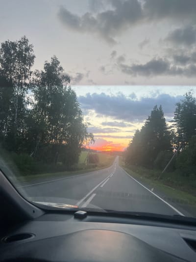 The image depicts a view from inside a car, looking out through the windshield at a rural road during sunset. The road curves to the left in the distance, with two lanes marked by white lines. A vibrant orange and yellow sunset is visible on the horizon, partially obscured by dark clouds and silhouetted trees. Dense evergreen trees line the right side of the road, while deciduous trees and fields are visible on the left. The foreground shows the car's dashboard and steering wheel, reflecting the colors of the sunset. The scene appears to be located in the vicinity of Kundravy, Russia, given the typical landscape and rural setting associated with the region. There are no people visible in the image.