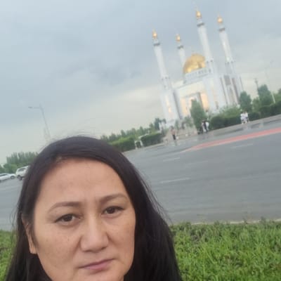 The image is a portrait of a woman taken outdoors. The woman has dark hair and appears to be looking towards the camera, with a slight smile. Her face is in the foreground and takes up a significant portion of the frame.
In the background, a large white building with golden domes and tall minarets is visible. This appears to be a mosque, possibly the Nur Gasyr Mosque in Aktobe, Kazakhstan, given the location context. The building is partially obscured by trees and greenery.
To the right of the building, a road with a red stripe is visible, separating it from a grassy area. There are some indistinct figures of people and vehicles on the road and near the building, suggesting it is an active public space.
The sky is overcast, indicating a cloudy or possibly rainy day. The lighting suggests it is daytime. The overall scene appears to be a view of a significant architectural landmark from a public area. The woman's expression is relaxed and content. There is no visible text in the image.