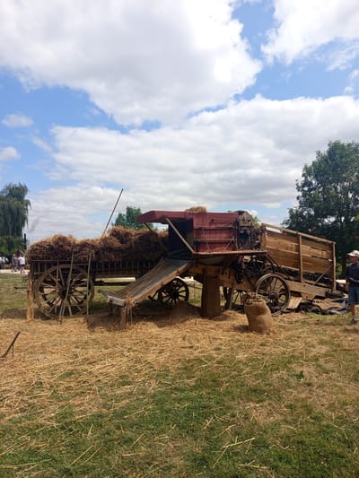 This image depicts an outdoor scene on a sunny day with a partly cloudy sky in Azannes-et-Soumazannes, France. The central focus is an antique threshing machine, a large piece of agricultural equipment, positioned in a field covered with dry grass and straw. The threshing machine is mounted on wooden wheels and appears to be made of wood and metal, with a red-topped structure. Hay is piled high on a wooden cart to the left of the threshing machine. In the background, several people are visible, some standing near a tall, weeping willow tree from which smoke is rising, suggesting a nearby building or activity. A person wearing a hat and shorts stands to the right of the threshing machine. Burlap sacks are placed on the ground near the equipment. The overall environment suggests a historical reenactment or demonstration of farming practices.