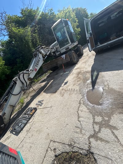 The image shows a construction site in what appears to be Silistra, Bulgaria, based on the Cyrillic lettering on the Terex excavator. A white van with its back door open is parked next to a Terex excavator. A puddle of water sits on the cracked concrete pavement. A set of tools is laid out beside the excavator. There are trees in the background and a blue sky.