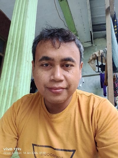 A man is posing for a selfie. He has short, dark hair and brown eyes. He is wearing a mustard-yellow t-shirt. Behind him is a green and white pillar and a clothesline with various items hanging on it. The lighting is natural, and the background suggests an outdoor or semi-outdoor setting. The bottom left of the image shows a watermark that indicates the photo was taken with a Vivo Y19s.