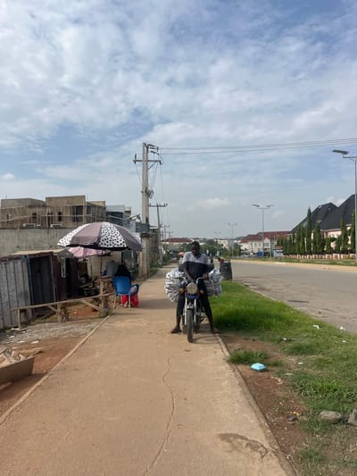 A man is seated on a motorcycle loaded with large bundles wrapped in plastic, parked on the side of a paved road in Karum, Nigeria. The motorcycle is on a dirt and grass median next to the road. The man is wearing a dark and gray t-shirt and dark pants. He is looking forward. The road has multiple lanes and is lined with neatly trimmed trees and modern houses with dark roofs. In the background, there are several houses under construction with scaffolding. On the left side of the image, there is a small roadside stall with a polka-dot umbrella providing shade over a table and some chairs. Electrical poles and wires are visible along the road. The sky is partly cloudy.