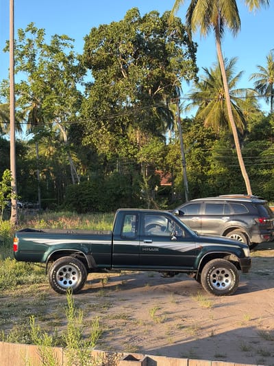 A dark green Toyota Hilux pickup truck is parked on a dirt lot. The truck is facing right and is in the foreground. The truck has a chrome trim along the side and a "HILUX" logo on the door. The truck also has off-road tires with "MONSTER TRUCK" written on the side. In the background, a dark gray SUV is parked to the right of the truck. Behind the vehicles, there is a dense jungle with palm trees and other greenery. The sky is clear and blue. The scene is bathed in the warm light of the late afternoon sun. The location is identified as Baan Taling Ngam, Thailand.