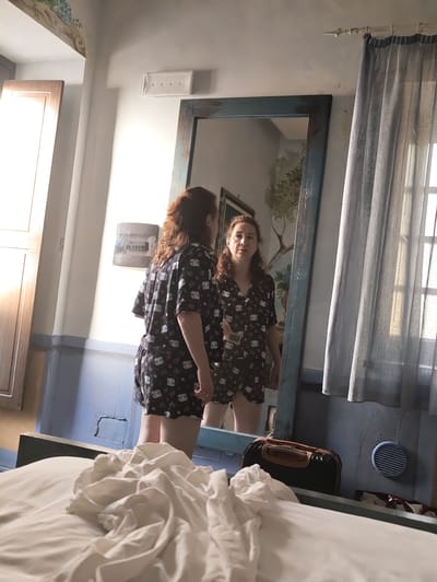 A woman with curly, auburn hair is standing in front of a large, ornate mirror, her reflection visible. She is wearing a black pajama set with a white cartoon print. The room appears to be a bedroom with a large bed in the foreground draped with white linens. A window with sheer grey curtains is visible to the right, allowing natural light to enter. A dark suitcase with tan accents rests on a surface below the mirror. A circular vent is mounted on the blue-grey wall behind the suitcase. The woman's hands are visible, and she appears to be adjusting her pajama shorts. The overall impression is one of a casual, domestic setting. The image provides no specific information about the city of Pozzuoli, Italy.
