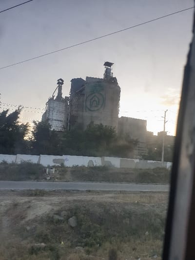 Two industrial concrete silos with green logos stand beside a road. The landscape appears dry and scrubby. A gray overcast sky serves as the backdrop. Wires and what seems to be part of a window frame are partially visible at the edges of the image, giving the impression of viewing the scene through a pane.