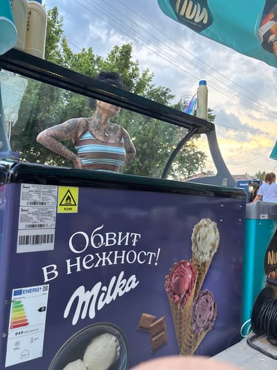 This image captures an outdoor scene at an ice cream stand in Kostievo, Bulgaria, during what appears to be late afternoon or early evening. The central focus is a Milka-branded ice cream freezer, primarily purple with white Cyrillic text reading "ОБВИТ В НЕЖНОСТ!" (Wrapped in tenderness!) and the "Milka" logo. The freezer displays images of various ice cream cones and scoops in a bowl, alongside Milka chocolate squares.
Affixed to the freezer are several labels: a technical data sheet from JUKA trade-Invest Ltd. for a "Display Freezer M400SL," indicating it is "Made in Ukraine" and uses R290 refrigerant, a flammable gas, as noted by a yellow warning sticker with a flame symbol. An energy efficiency label shows a "D" rating, 949 kWh/annum, and -23°C operating temperature.
Reflected in the glass top of the ice cream cart is a person, likely a woman, with dark hair and extensive tattoos covering her arms, shoulders, and chest. She wears a brown and light blue striped tank top and has her hands on her hips, facing to the right in profile. On the top surface of the cart, several items are visible, including light-colored bottles (one with a blue cap) and clear glasses containing colorful elements, possibly straws.
In the background, on the far right, a second person with reddish-brown hair tied back, wearing a white t-shirt, is partially visible, facing away from the camera. The setting includes green trees, overhead power lines, and a cloudy sky suggesting approaching dusk. Part of a light blue awning or flag, bearing reversed text that includes "ICE CREAM ADVENTURE," is visible in the upper right. A coiled black cable or hose is present in the lower right foreground. Distant structures, including one with "Nudel" visible on its side, are vaguely discernible.