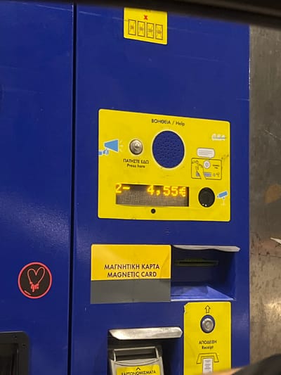 A close-up photograph depicts a dark blue automated payment kiosk, likely for tickets or services, situated within the Municipal Unit of Thebes, Greece.
The machine's upper section features a small yellow sticker with a red 'X' overlaid on four crossed-out square icons, indicating unavailable functions. The central area is dominated by a large yellow panel containing interactive elements. At the top right of this panel, text reads "ΒΟΗΘΕΙΑ / Help". A silver circular button with a loudspeaker icon and "ΠΑΤΗΣΤΕ ΕΔΩ / Press here" below it is present, adjacent to a circular speaker grille. An amber-lit digital display shows "2- 4.55€". To the right of the display, a small black circular lens or sensor is visible, alongside graphic instructions for inserting a card and a security camera icon.
Below the central panel, a yellow rectangular sign indicates "ΜΑΓΝΗΤΙΚΗ ΚΑΡΤΑ / MAGNETIC CARD", positioned above a recessed slot for magnetic card input. To the left, a circular black sticker features a stylized pink heart with a dripping effect. At the bottom right, another yellow panel displays an upward arrow icon, a circular button, and "ΑΠΟΔΕΙΞΗ / Receipt" text, indicating a slot for dispensing receipts. Directly below the magnetic card slot, a metal tray and a partially visible yellow label with "ΧΑΡΤΟΝΟΜΙΣΜΑΤΑ" (Banknotes) suggest a cash payment or change dispensing mechanism. No individuals are clearly visible, though a hand or sleeve is partially present at the bottom right edge of the frame, near the receipt dispenser. The background beyond the machine appears dark and indistinct.