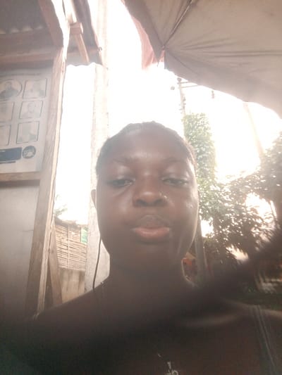 The image is a close-up selfie of a Black woman with braided hair. The background is overexposed, but details suggest an outdoor environment in Mbribit, Nigeria. To the left, a wooden structure displays framed portraits. Behind the woman, there is some foliage and what appears to be a makeshift fence or building. Above, there is a faded awning or umbrella.